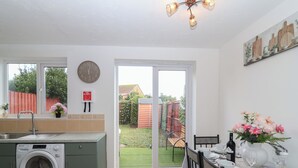 A compact kitchen with a washing machine, a dining area with a table set for two, and a large sliding glass door leading to a garden. - The Cosy View (Paignton)