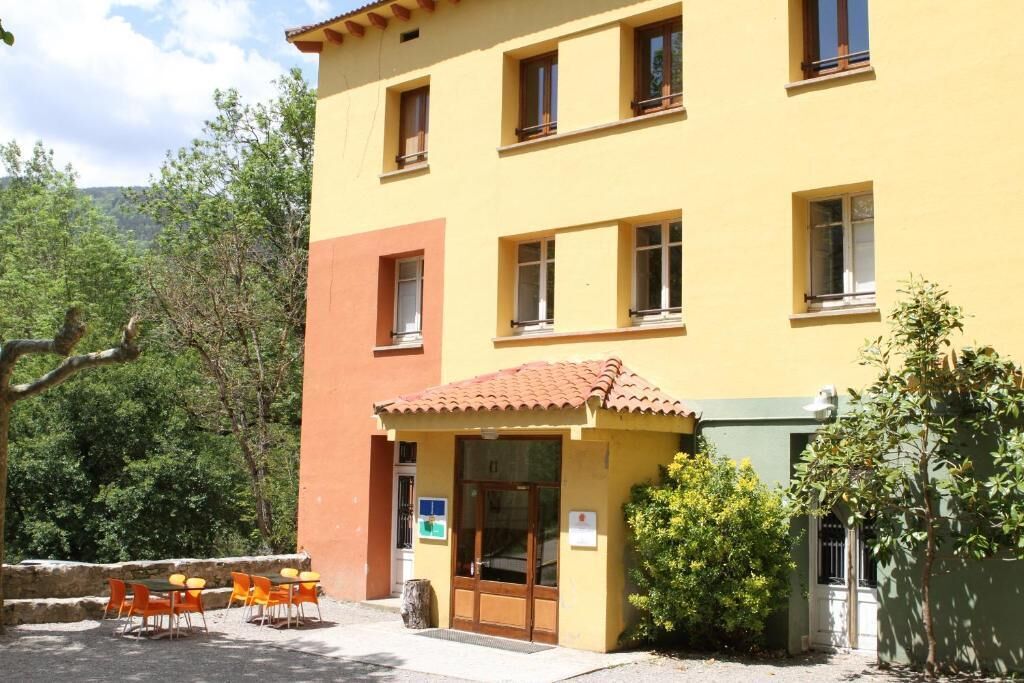 A yellow building with a red-tiled entrance, outdoor seating, and greenery around.