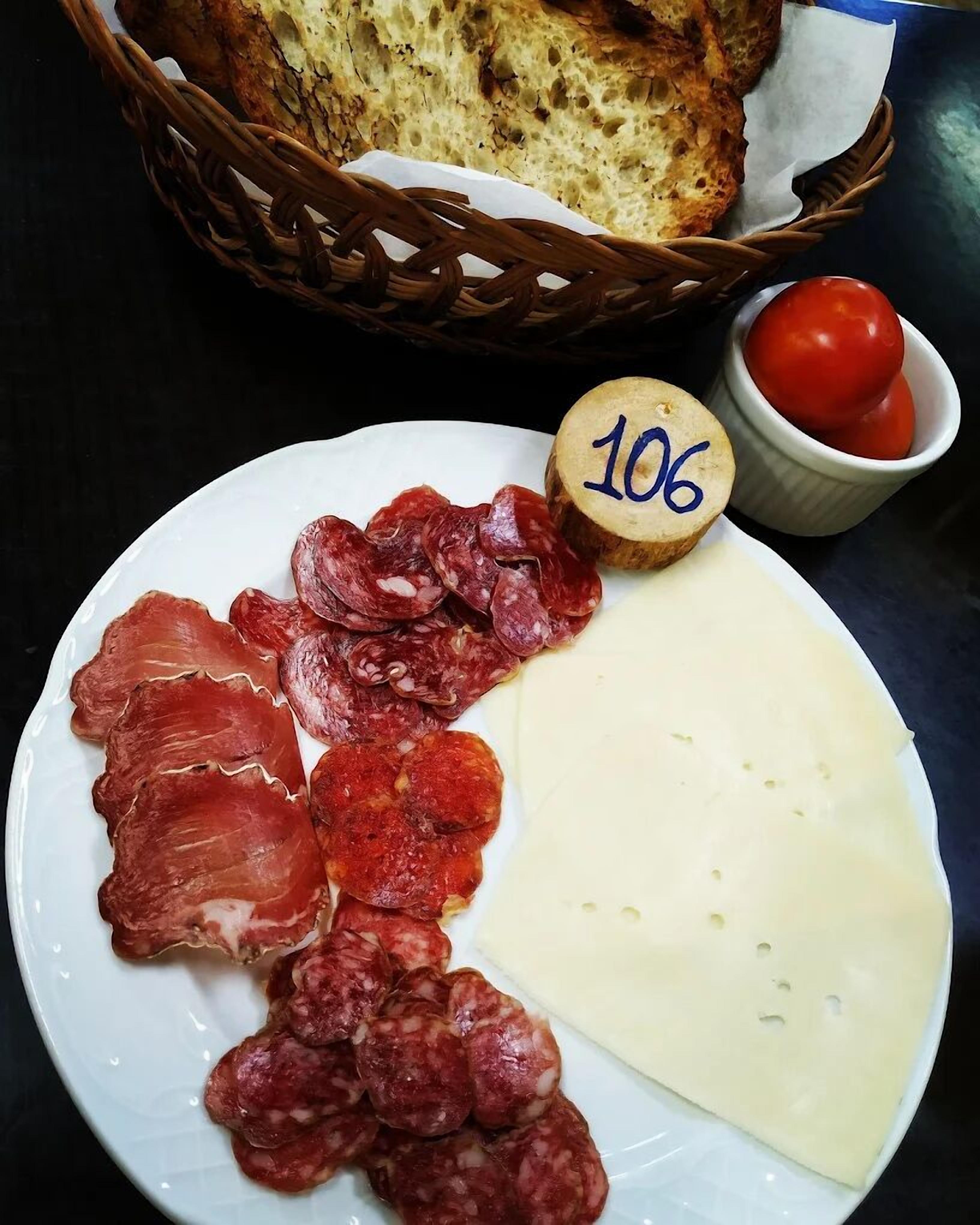 A plate with various cured meats and cheese, a wooden number marker, a basket of bread, and a small bowl of tomatoes.