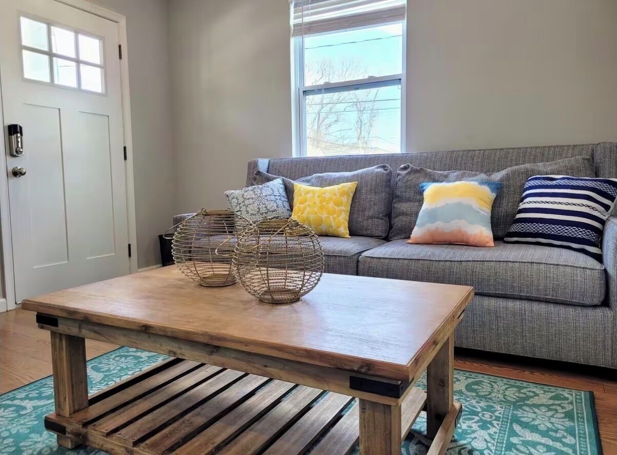 Ein Wohnzimmer mit Couchtisch aus Holz, einem grauen Sofa mit bunten Kissen und einem Fenster mit Jalousien.