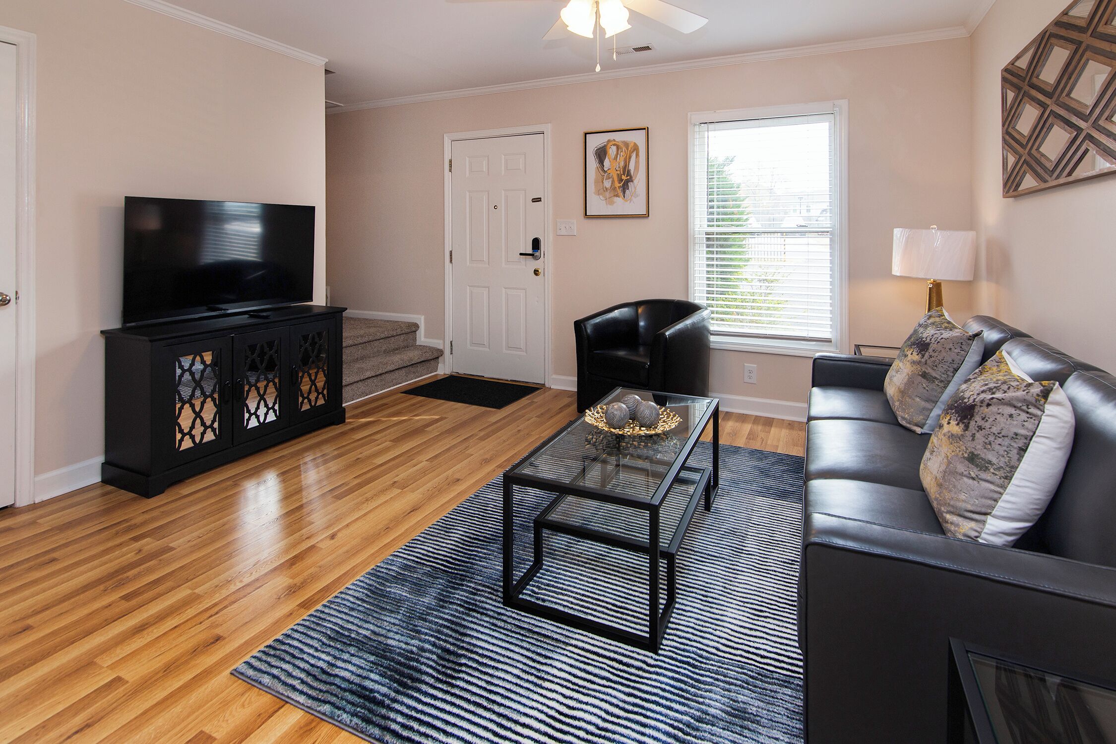 A living room with a flat-screen TV, a glass coffee table, a black leather sofa, and a patterned rug.