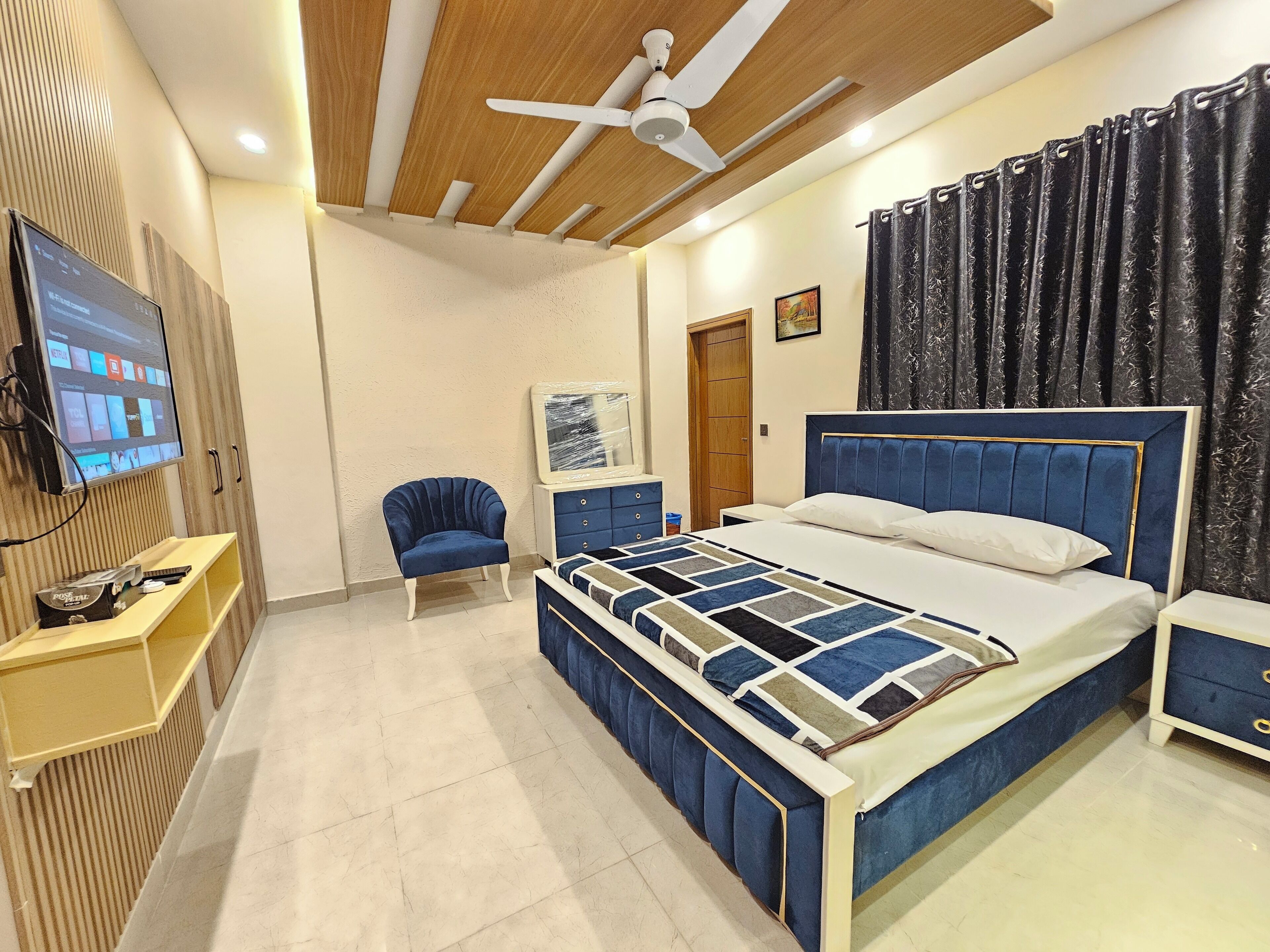 A modern bedroom with a large bed, a TV mounted on the wall, a chair, and a wooden ceiling.