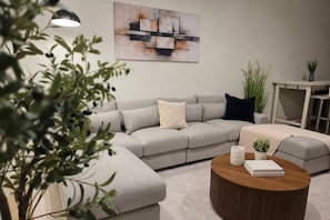 Una sala de estar moderna con un sofá seccional gris, una mesa de centro redonda de madera y un cuadro abstracto minimalista en la pared.