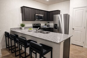 A kitchen with dark cabinets, a stainless steel refrigerator, a microwave, a granite countertop, and black bar stools.