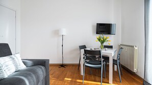 Ein modernes Wohnzimmer mit einem grauen Sofa, einem Esstisch mit Stühlen, einer Stehlampe und einem an der Wand montierten Flachbildfernseher.