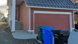 A two-story house with a garage, a red exterior, and a grey roof. There are three trash bins in front of the garage. - Unwind in this serene environment with close proximity to the airport. (Calgary)