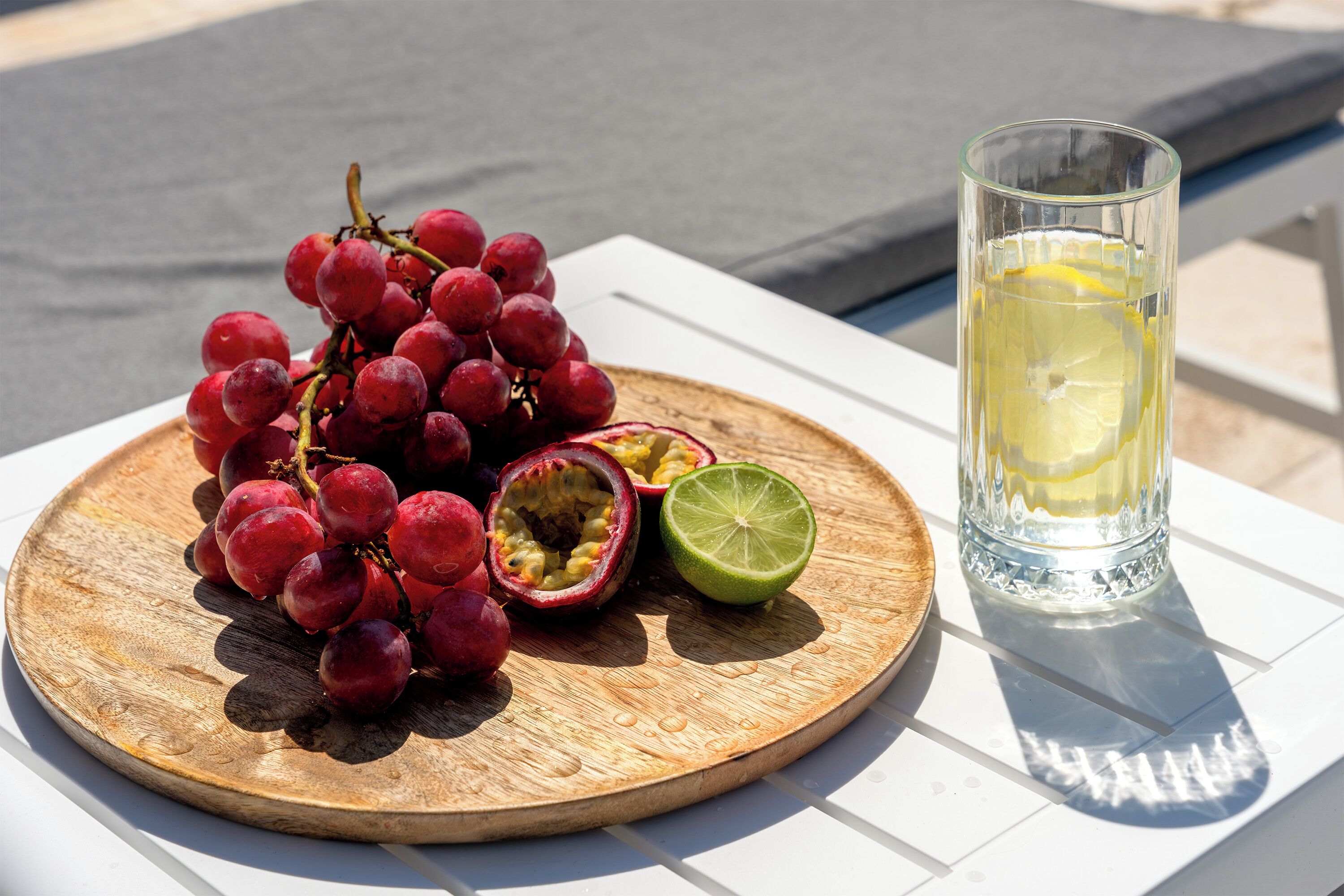 Une planche en bois sur laquelle on a disposé des raisins rouges, une moitié de fruit de la passion et un verre de limonade, le tout sur une table blanche.