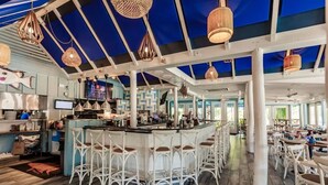 Een ruim restaurant met een hoge plafond, zichtbare balken en een bar met witte barkrukken.