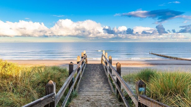 Een houten looppad leidt naar een strand met een helder uitzicht op de oceaan en een houten pier die het water in reikt.