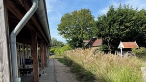 Een overdekt buitengebied met een grindpad, houten constructies en een gazon met hoge planten.