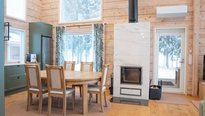 A cozy cabin interior with a wooden dining table, chairs, a built-in stove, and large windows offering a view of the forest. - Villa Siltasaari (Sotkamo)