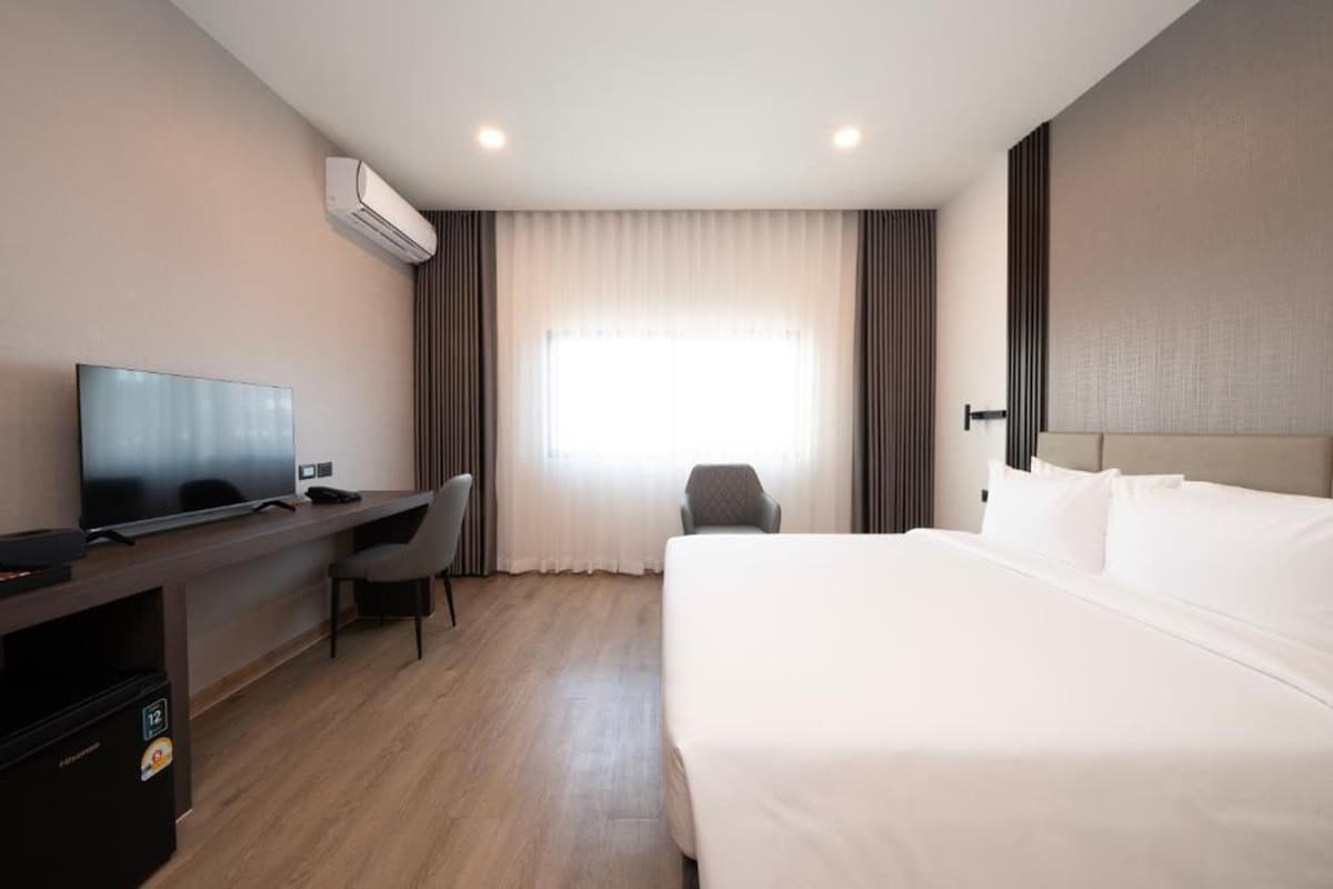 A hotel room with a bed, a desk with a television, a chair, and an air conditioning unit.