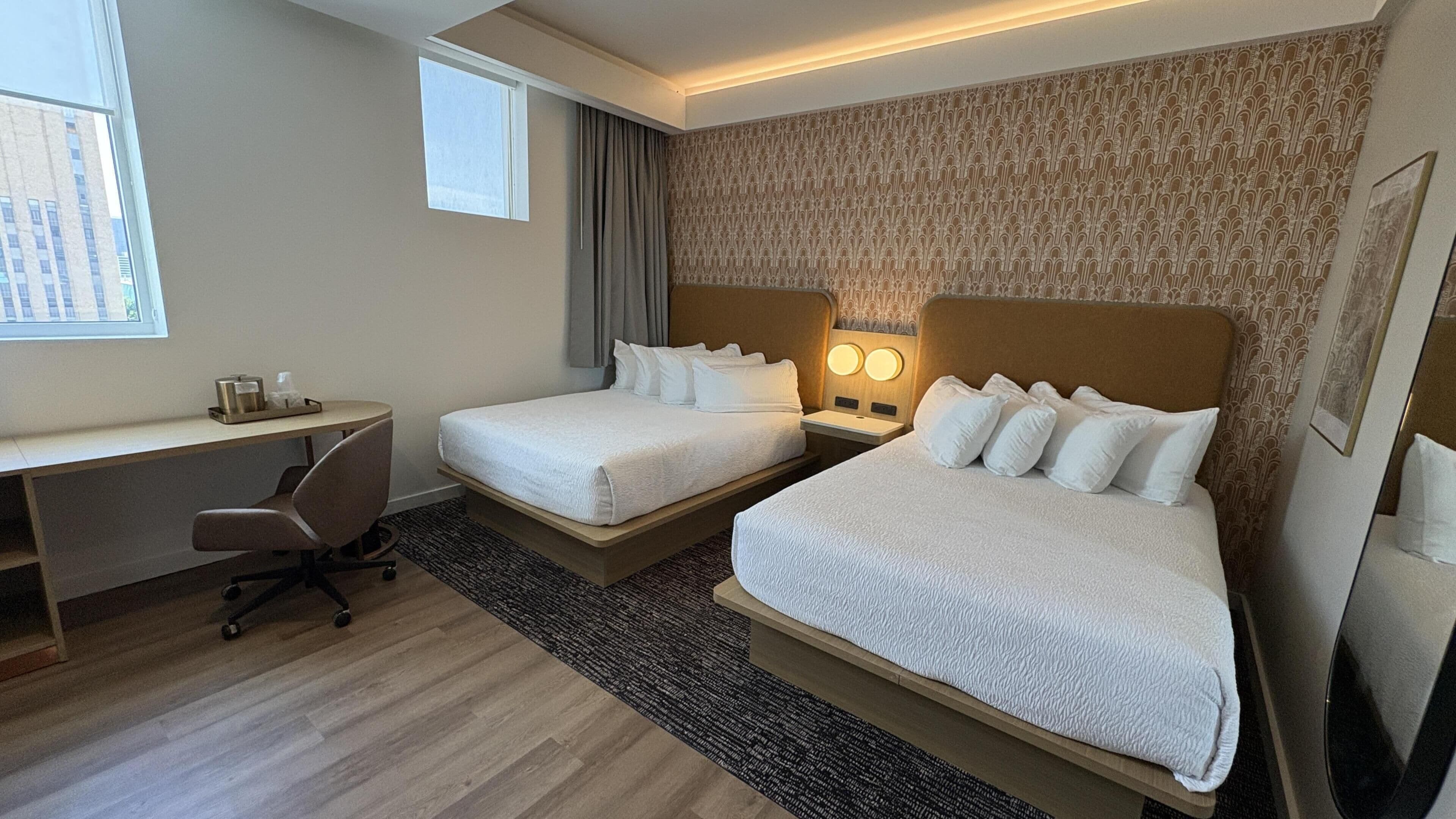 A hotel room with two beds, a desk, a chair, and a window.