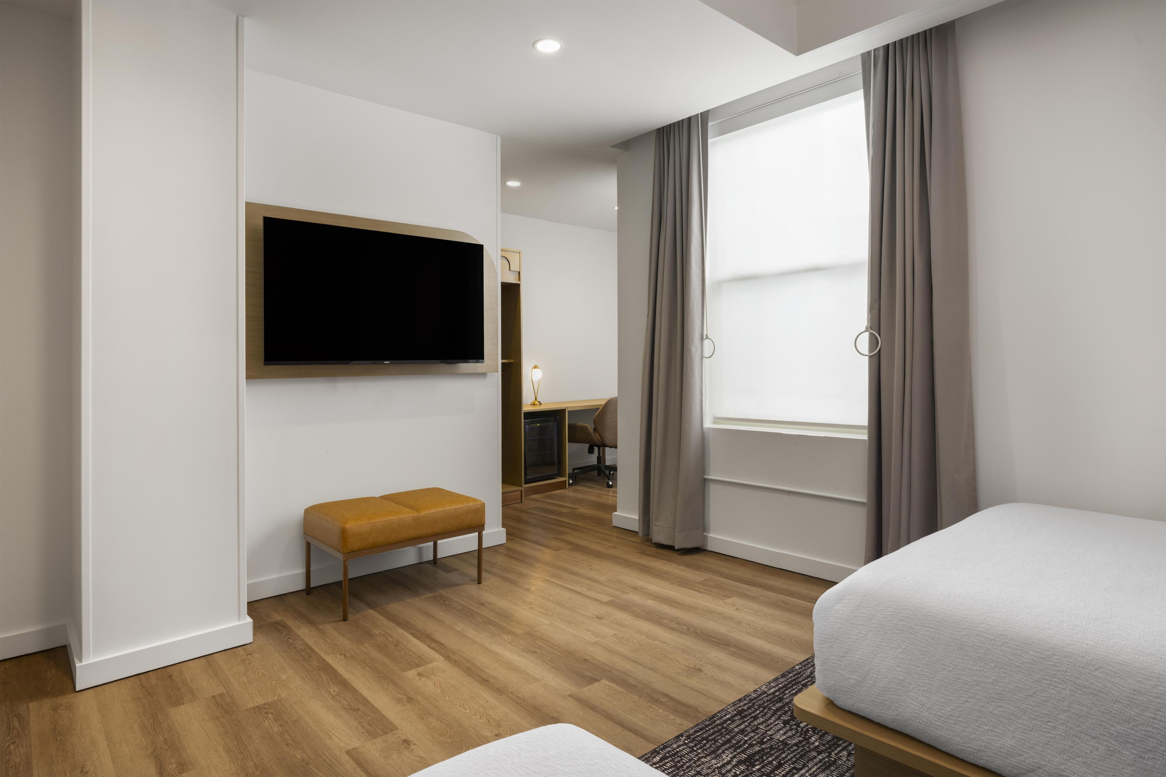 A hotel room with a flat-screen TV mounted on the wall, a wooden bench, a desk with a chair, and a large window with curtains.