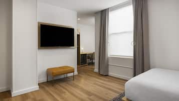 A hotel room with a flat-screen TV mounted on the wall, a wooden bench, a desk with a chair, and a large window with curtains.