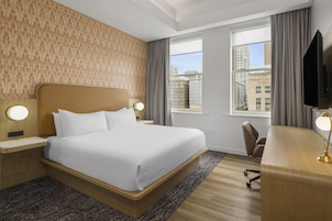 A hotel room with a large bed, a desk with a chair, a TV, and a window with a city view.