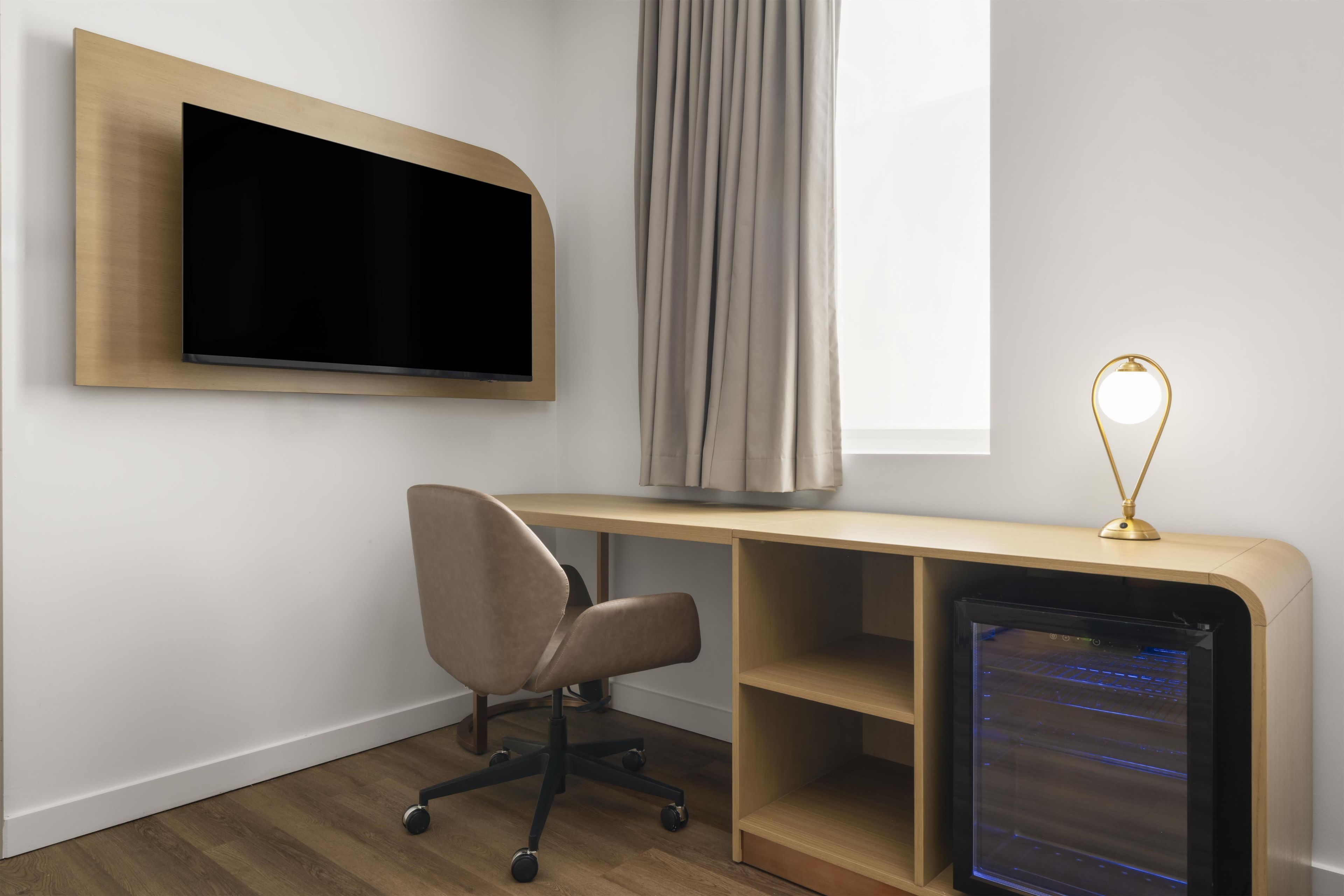 A modern room with a flat-screen TV mounted on the wall, a desk with a computer, a chair, and a lamp.