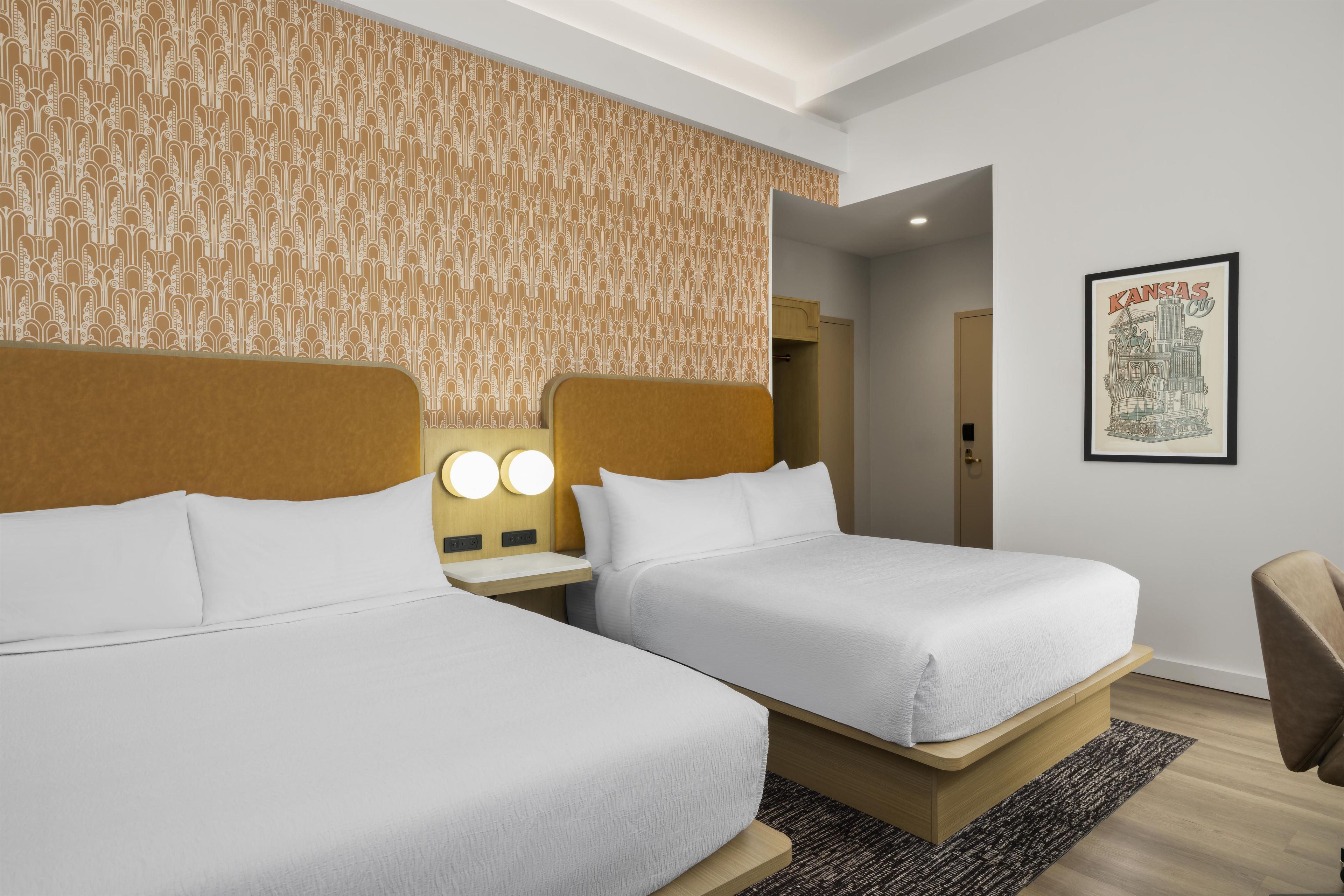 Quarto de hotel com duas camas, cabeceira com luminária e uma imagem emoldurada de vista urbana na parede, além de piso de madeira.