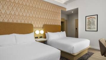 Quarto de hotel com duas camas, cabeceira com luminária e uma imagem emoldurada de vista urbana na parede, além de piso de madeira.