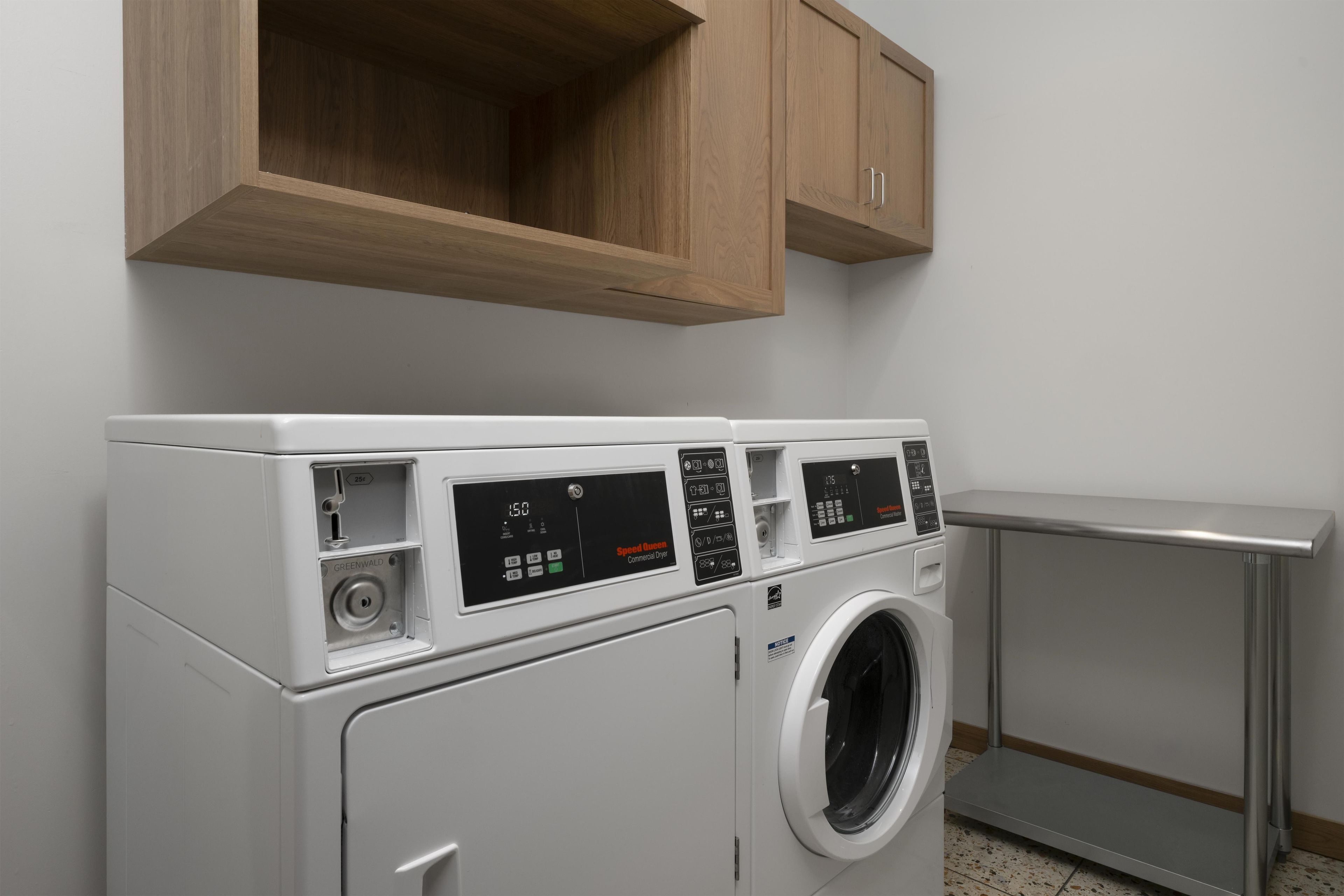 A front-loading white washer and dryer unit with a digital control panel, a wooden cabinet above, and a metal shelf to the right.