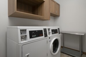 A front-loading white washer and dryer unit with a digital control panel, a wooden cabinet above, and a metal shelf to the right. - Aiden By Best Western Kansas City Downtown (Kansas City)
