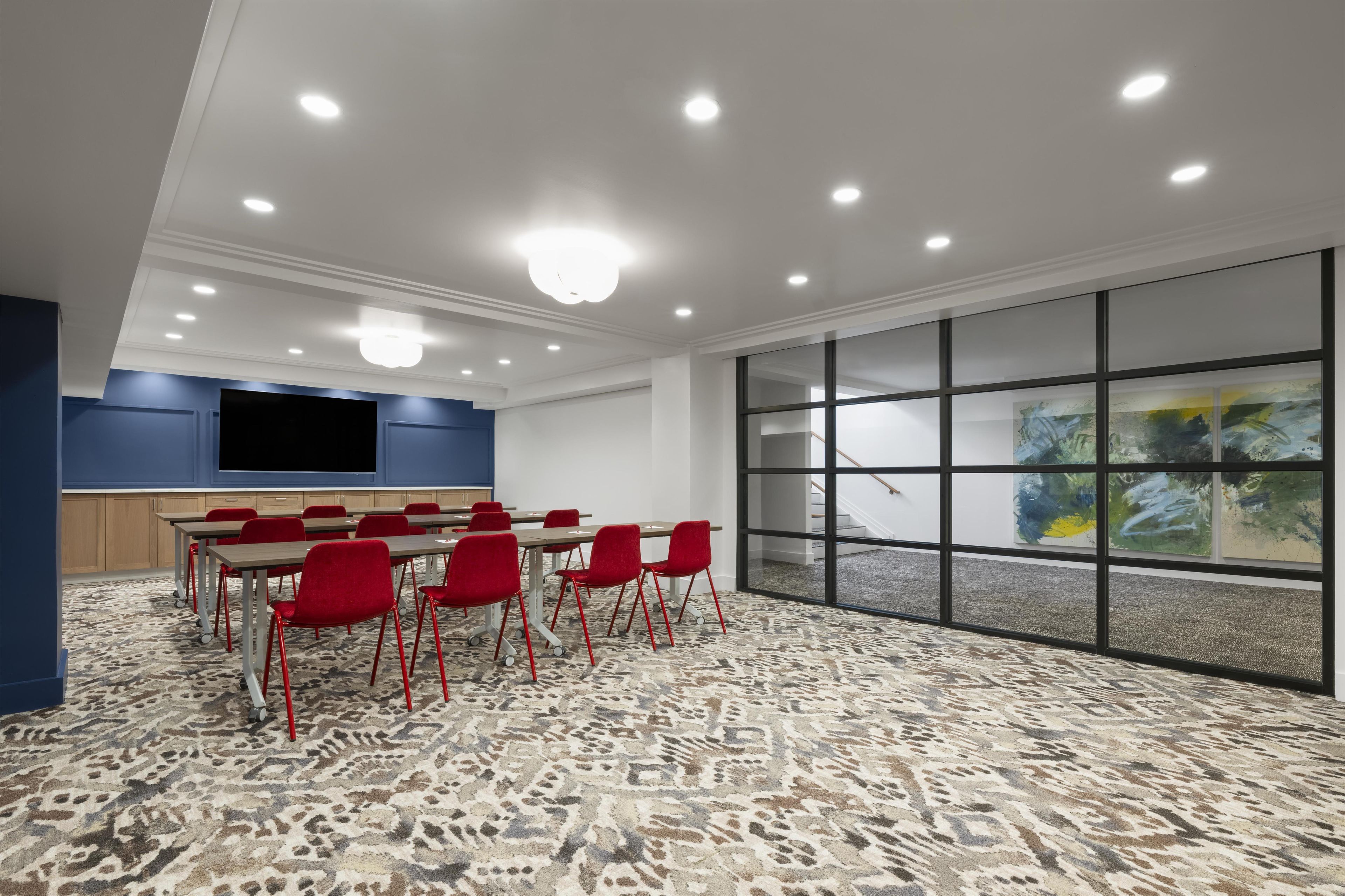 A conference room with a large table, red chairs, a flat-screen TV, and a view through a glass wall.