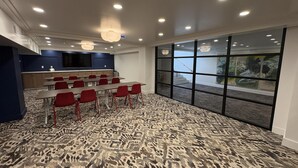 A conference room with a patterned carpet, red chairs, a long table, a large screen, and a view through a glass wall.