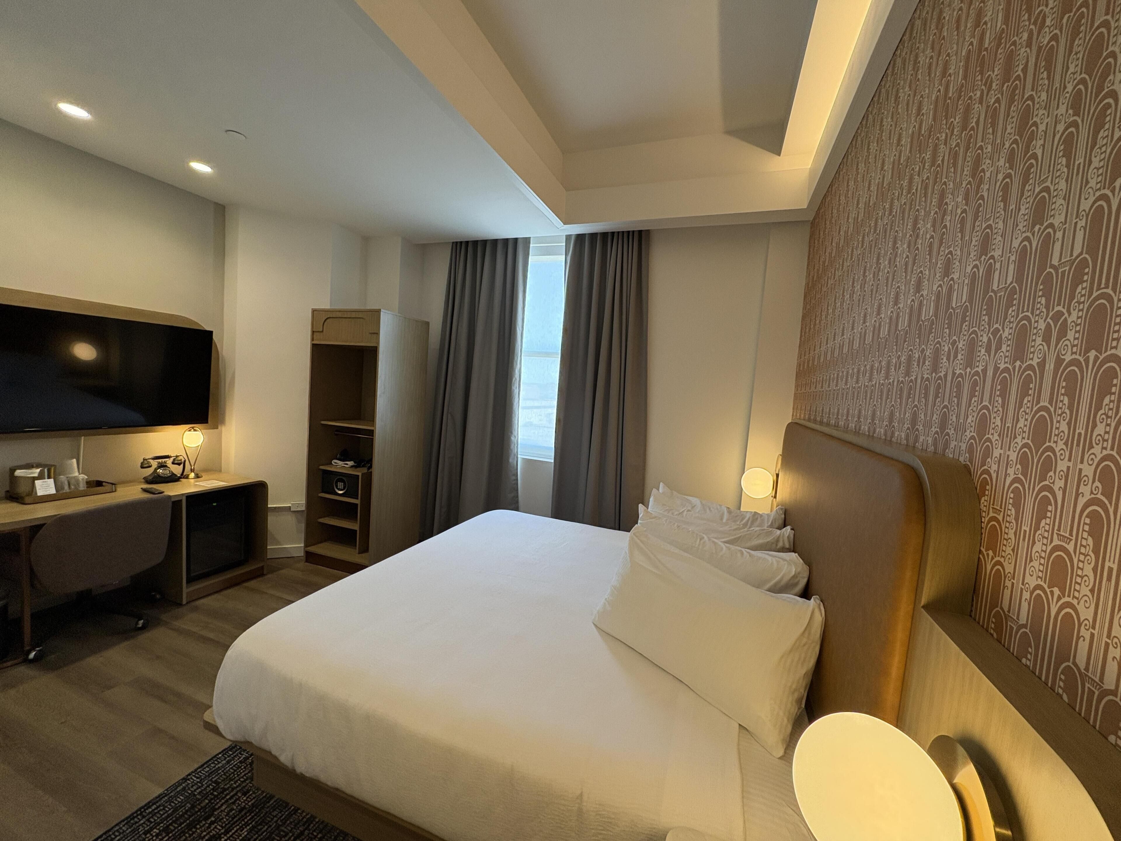 A hotel room with a bed, a desk with a TV, a wardrobe, and a window with curtains.