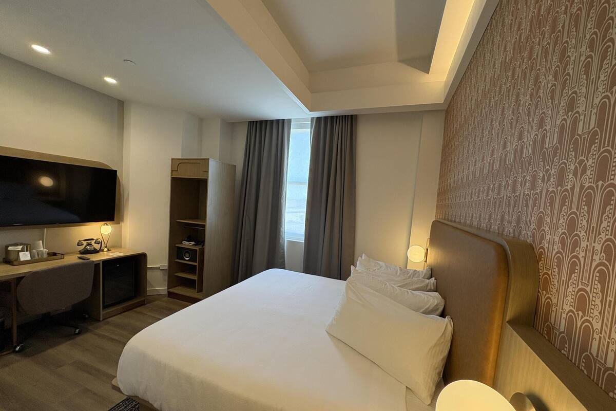 Une chambre d’hôtel comprenant un lit, un bureau avec une télévision, une armoire et une fenêtre avec des rideaux.