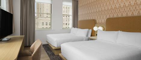 A hotel room with two beds, a desk, a chair, a TV, and a view of buildings through the windows.