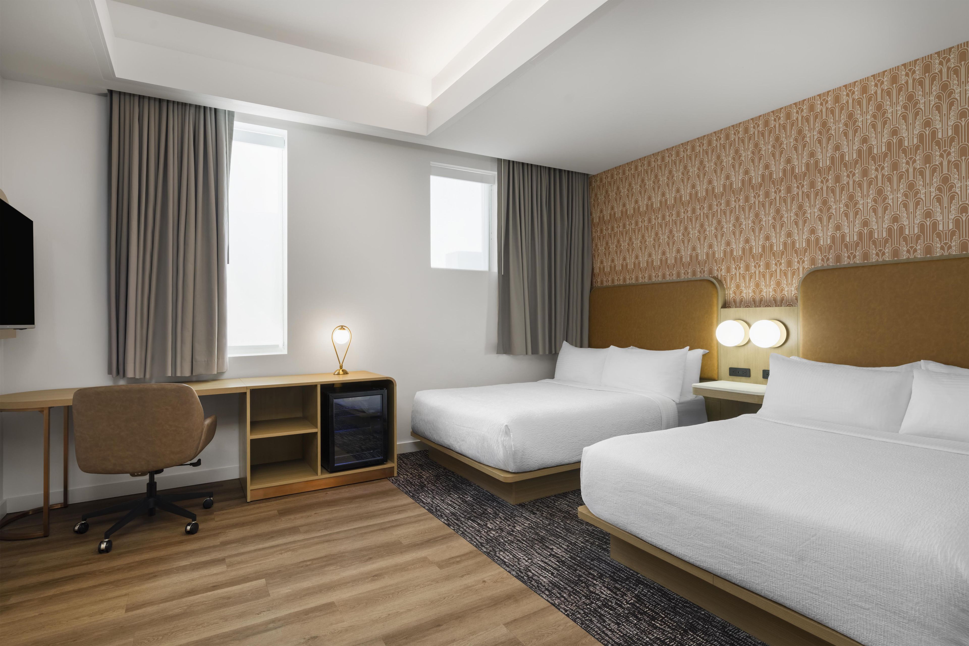 A hotel room with two beds, a desk, a chair, a TV, and a patterned wall.