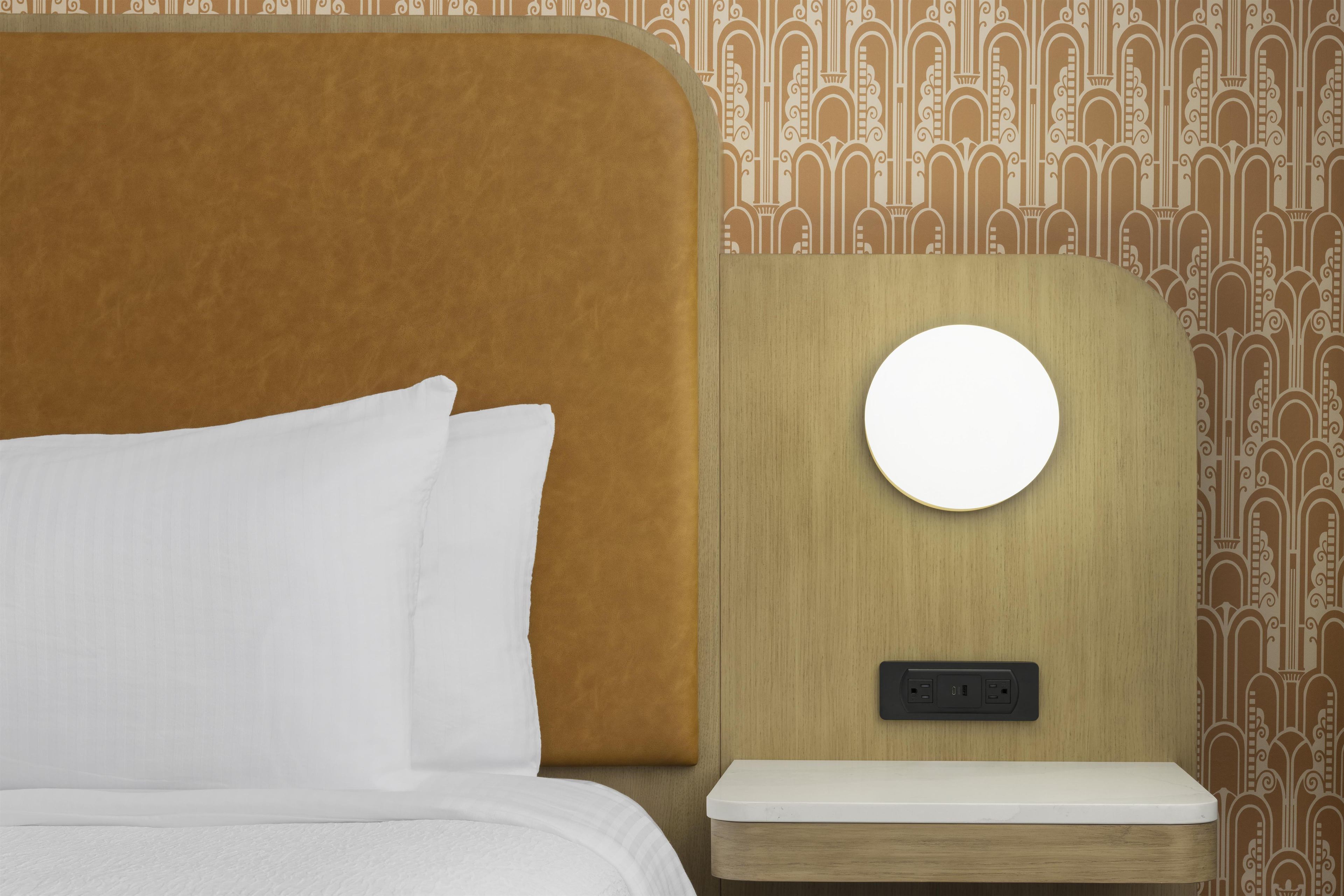 A bed with white bedding, a round mirror on a wooden nightstand, and a wall with a patterned wallpaper.