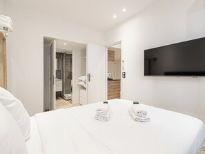 Una camera d'albergo con un letto, due asciugamani arrotolati, una TV e l'area della doccia visibile attraverso una porta aperta.