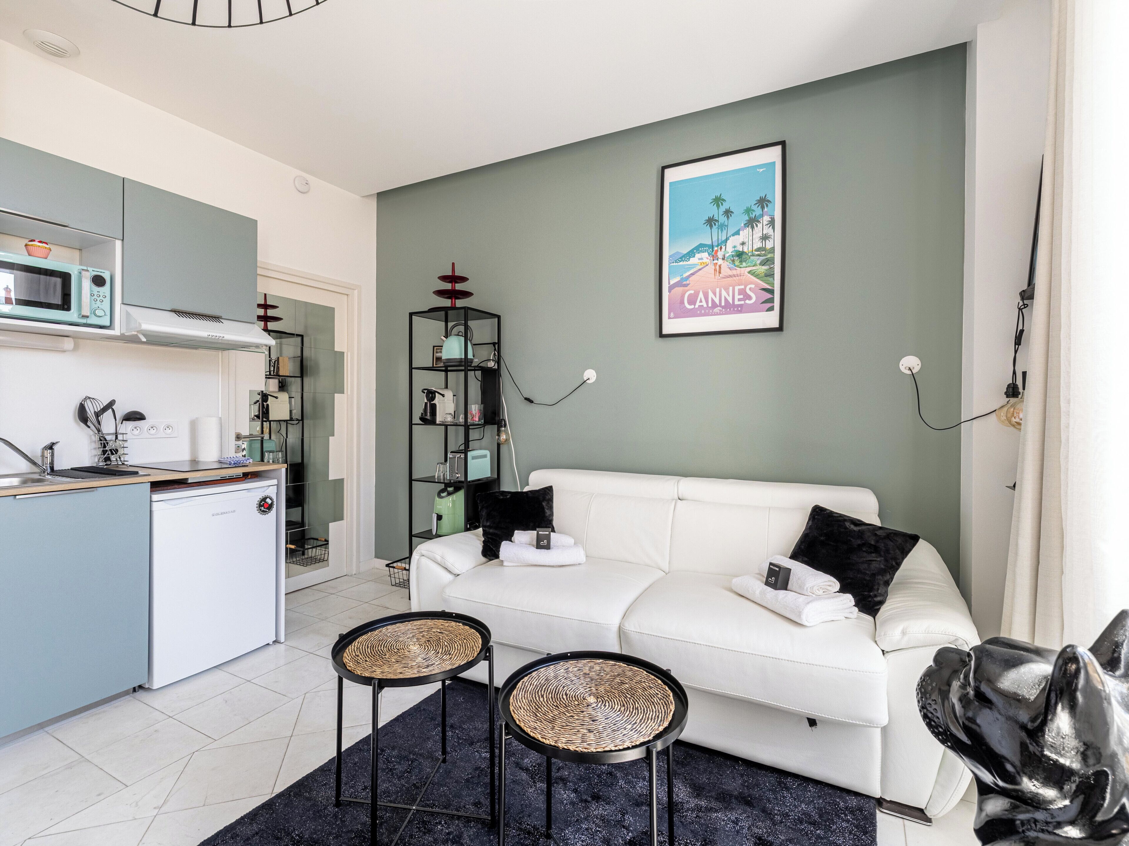 Ett modernt vardagsrum med en vit soffa, två flätade sidobord, en köksdel med mikrovågsugn och en inramad tavla med ett motiv från Cannes på väggen.
