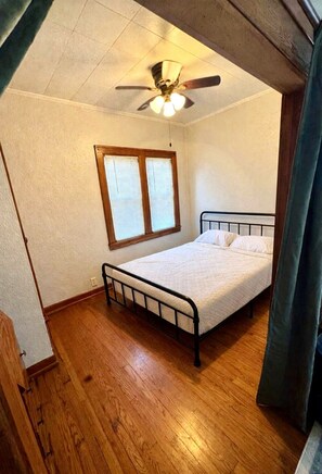 A small, single-bed room with a wooden floor, a ceiling fan, and a window with blinds. - Cozy Duplex in the Heart of Benson (Omaha)