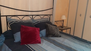 A bed with a metal frame, multiple pillows, and a striped bedspread. There is a small table with a lamp beside the bed. - "Le Pesquit" large, fully-equipped 36m2 studio on one level: private terrace. (Lescar)