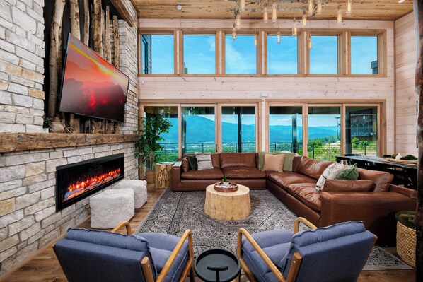 A spacious living room with a stone fireplace, leather sofas, a large TV, and a view of mountains through large windows. - MODERN NEW CONSTRUCTION! Naturehaus Family Retreat–Pool, Golf, Pickleball, Views (Pigeon Forge)