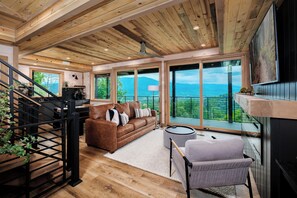 A spacious living area with a wooden ceiling, large windows offering a mountain view, a brown sofa, and a round table. - MODERN NEW CONSTRUCTION! Naturehaus Family Retreat–Pool, Golf, Pickleball, Views (Pigeon Forge)