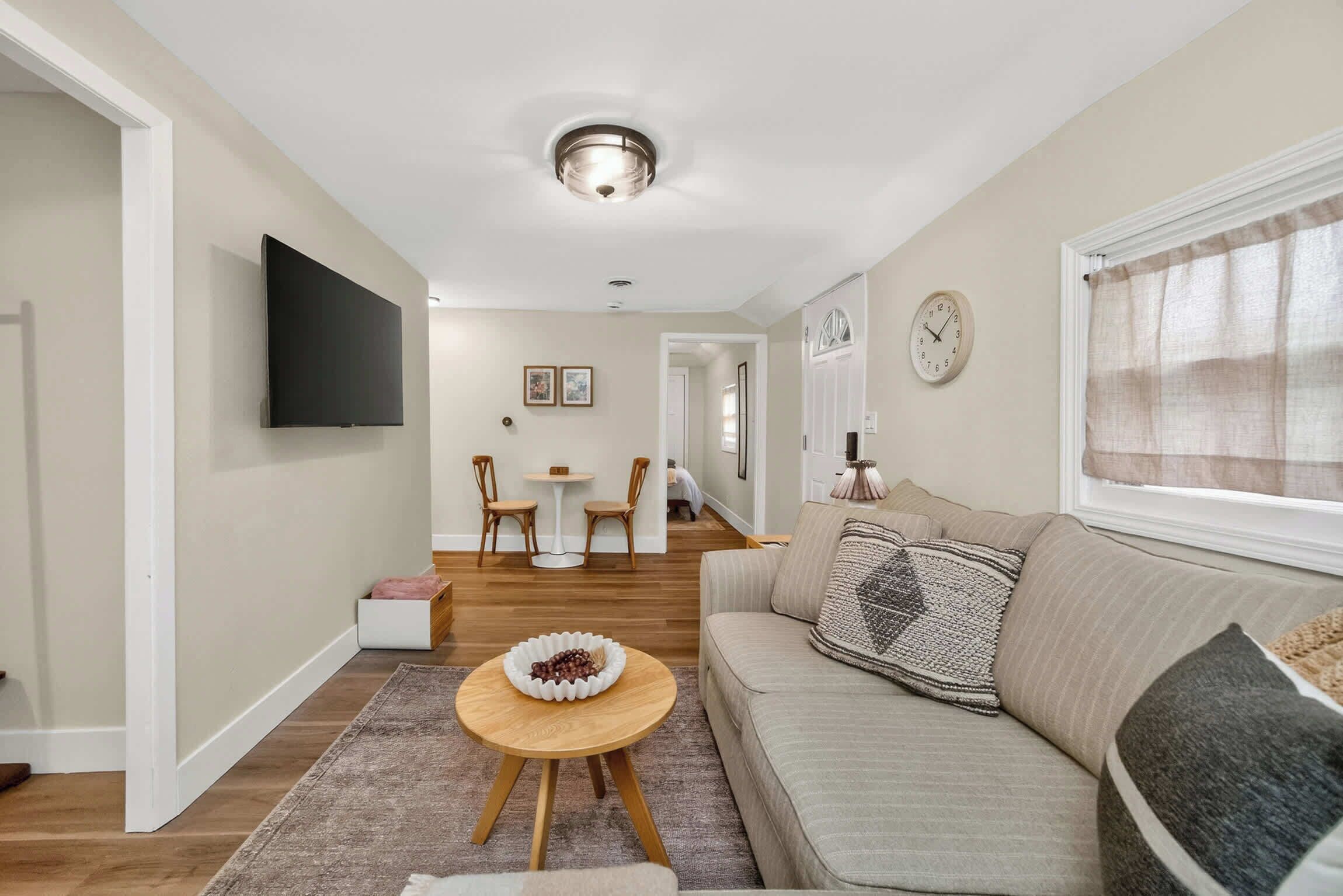 Een moderne woonkamer met een bank, een salontafel en een flatscreen-televisie aan de muur. Er is een eethoek met een tafel en stoelen, en een gang die naar een andere kamer leidt.