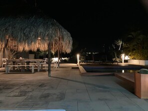 Un espace extérieur aménagé pour la nuit, avec une piscine, des chaises longues et une structure dotée d’un toit de chaume.