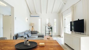 Ett modernt vardagsrum med ett matbord i trä, en grå soffa, en tv på ett vitt skåp och ett fönster med persienner.