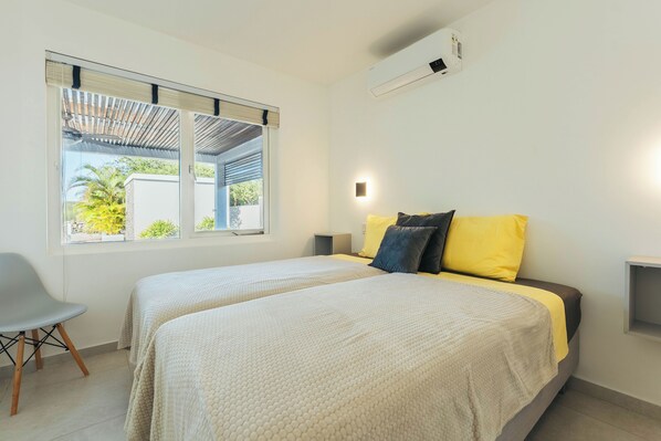 Une chambre avec deux lits, une fenêtre avec des stores, une chaise et un climatiseur fixé au mur.