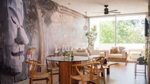 Ein modernes Wohnzimmer mit einem runden Glastisch, umgeben von Holzstühlen, einem großen abstrakten Wandbild und einem Fenster mit Blick auf Grünflächen.