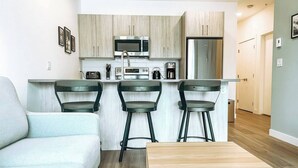 Cozinha moderna com micro-ondas, cafeteira e frigorífico. Há três bancos altos e uma mesa de madeira.