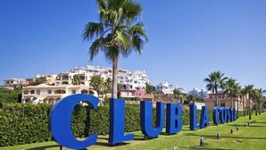 Une enseigne avec le texte « CLUB LA COSTA » en grandes lettres bleues, placée sur un fond représentant une ville côtière avec des bâtiments blancs et des palmiers.
