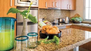 Une cuisine avec des meubles en bois, un four en acier inoxydable, un four à micro-ondes, un plan de travail en granit, un bol de croissants, une plante verte et un pichet contenant un liquide vert.