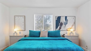 Un dormitorio con una cama grande, dos mesitas de noche con lámparas, un ventanal con persianas y dos cuadros enmarcados en la pared.