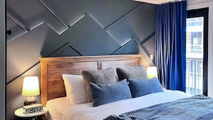 A modern bedroom with a wooden headboard, a bed with white and blue bedding, two bedside lamps, and a view of the city through a window with blue curtains. - The Nook Hydeaway at Ascent (Canmore)