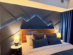Een moderne hotelkamer met een geometrisch plafondontwerp, een houten hoofdbord en een net opgemaakt bed met meerdere kussens.