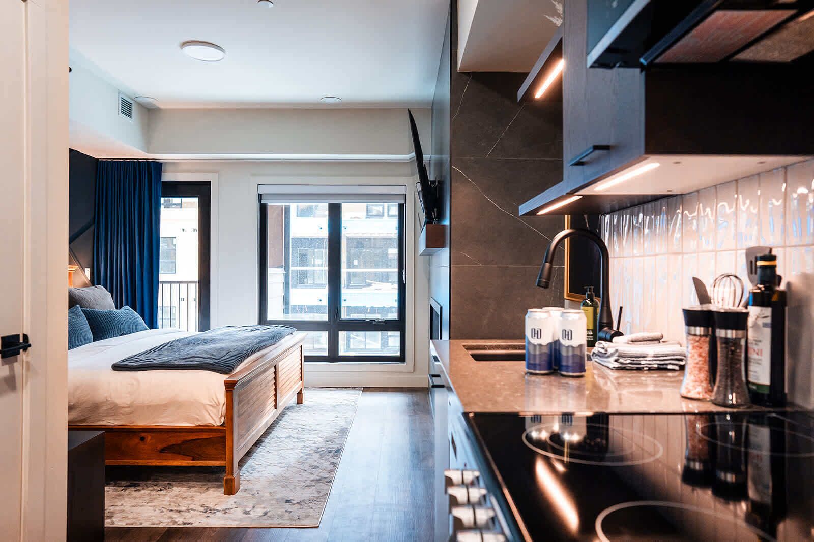 A modern hotel room with a bed, a large window, a kitchen area, and a view of the city.